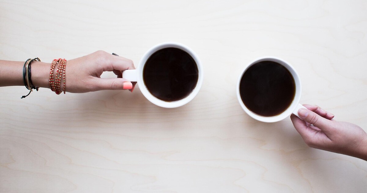 Zwei Kaffeetassen mit zwei Händen gegenüber am Tisch
